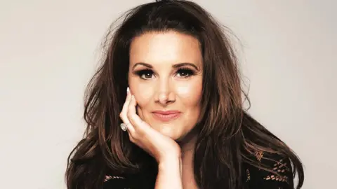 Sam Bailey Sam Bailey looking at the camera with her hand on her chin.