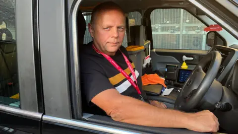 Gem O'Reilly/BBC Brian McDonough in a black t-shirt in his private hire vehicle. His driver's side window is open and he is resting his arm on the window edge. 