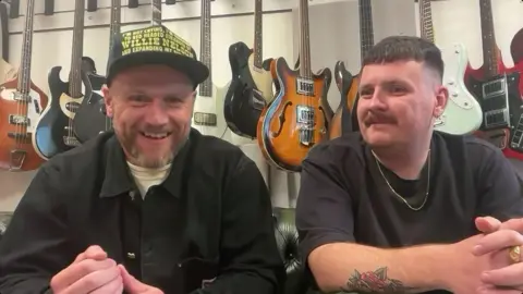 Two men are looking at the camera. The man on the left is wearing a white t-shirt and black jacket, has a beard and is wearing a cap. The man on the left is wearing a black t-shirt, has short, dark hair, he has a gold chain around his neck, and has a tattoo of roses on his right arm