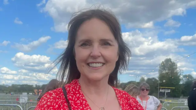 Gill Melder has short brown hair and is smiling while wearing a red dress with white detailing. She is standing in front of small metal barriers.