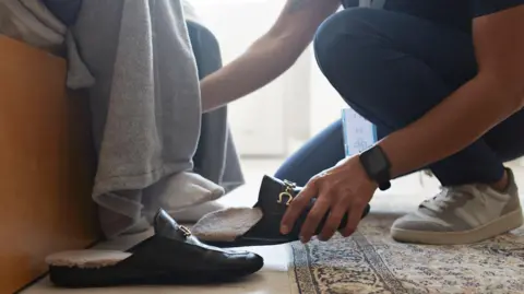 Getty Images A close up of a healthcare professional in dark blue scrubs and wearing a black watch, crouched down on a carpet and helping someone's feet into a pair of black shoes.