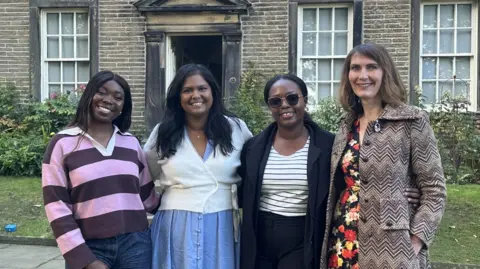 Olivia Courtney-Ashton/BBC Four women stand outside the Bronte Parsonage an old stone house. They are standing arm in arm. 