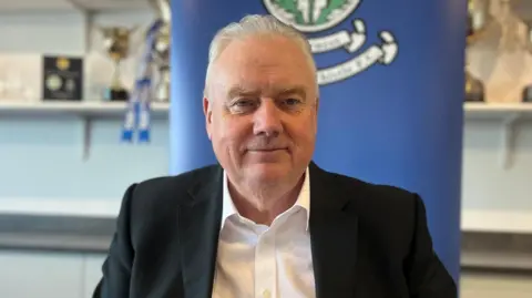 Alan Savage is sitting in the boardroom at Inverness Caledonian Thistle's stadium. He is an older man with white hair and clean shaven. He is wearing a black suit jacket over a white shirt.