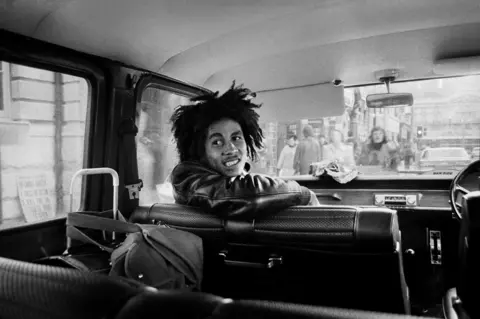 Dennis Morris Black and white photograph of Bob Marley in the passenger seat of a van with his body partially turned towards the back of the vehicle