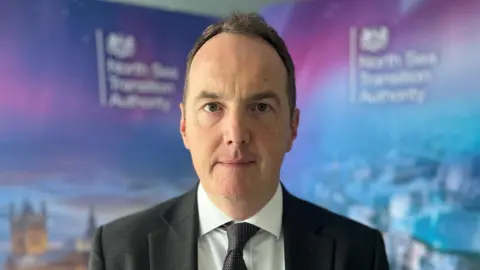 BBC A man with brown hair wearing a dark tie, white shirt and dark suit stands in front of a blue wall with the North Sea Transition Authority's logos.