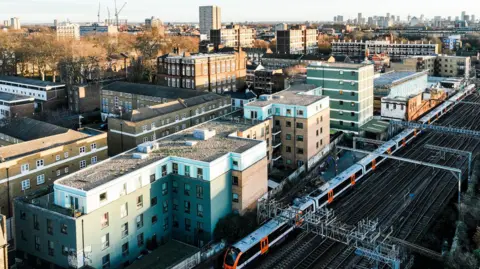 Getty Images A train line runs past a residential area in London, with the London skyline in the background. The residential area mostly consists of low-rise blocks of flats. A train is running on the train tracks past the flats.