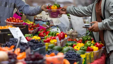 Getty Images A trader hands over a punnet of vegetables to a customer on a market stall, filled with fruit and vegetables.