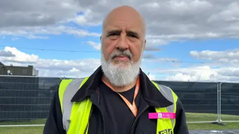 Shivani Chaudhari/BBC Rhys Hughes looking at the camera while outside at the festival site. He has a bald head and a long grey beard and moustache. He is wearing a hi-vis jacket and a lanyard and there is fencing behind him.