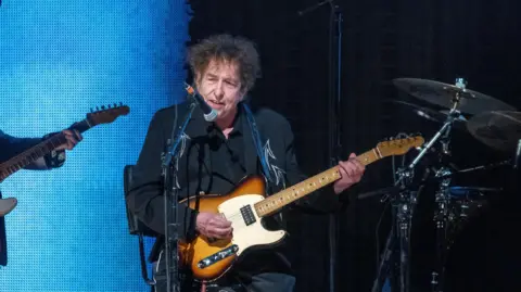 Getty Images Bob Dylan playing a sunburst guitar while dressed in black, sitting on a stool