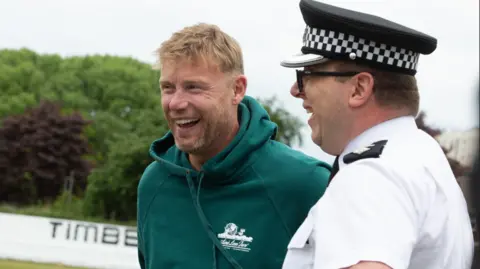 Merseyside Police Freddie Flintoff, wearing a green hoodie, laughs as he shares a joke with a police officer on the cricket pitch