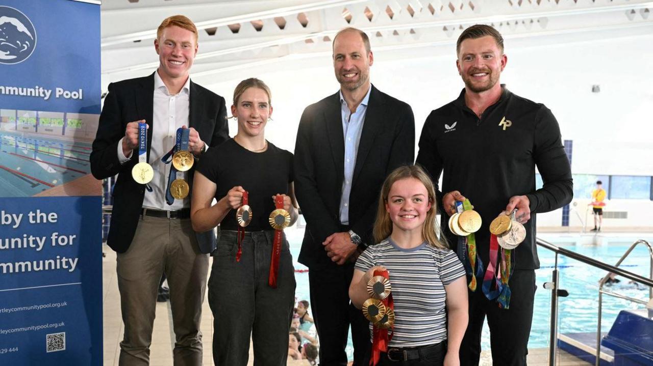 Prince William standing inside Birtley Community Pool. He is joined by Tom Dean, Olympian Adam Peaty and Paralympians Maisie Summers-Newton and Louise Fiddes. The swimmers are holding a mix of gold, silver and bronze Olympic medals.