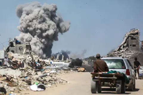 AFP A Palestinian man sits on a trailer behind a car on a road with smoke billowing and destroyed buildings in the background, following an Israeli strike in Jabalia in northern Gaza in May.