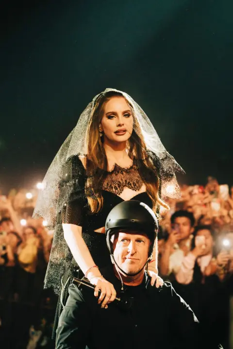 Kirby Gladstein Lana Del Ray in what looks like black bridal gear sits on the shoulders of a man in a black helmet and jacket as crowds photograph and film them on mobile phones.