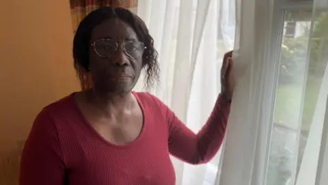 BBC Eslyn Daley in her living room. She is wearing glasses and a red scoop-necked top. She is near a window and has her hand leaning on the window frame. which has net curtains at it. We can see there is a garden behind her with some grass and other greenery. 