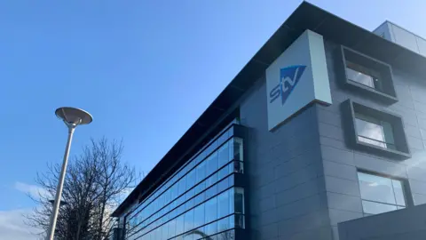 Exterior view of the STV building with modern glass and metal façade, featuring the STV logo on the corner. A row of leafless trees and a streetlamp line the pavement in front, with a clear blue sky overhead.