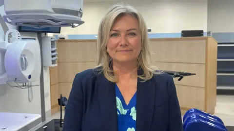 Jessica Bayley/BBC A smiling woman stood in front of a white piece of hospital equipment. She has shoulder-length, blonde hair and is wearing a blue suit jacket with a blue top underneath which has a floral pattern on. 