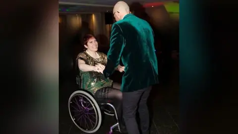 Family photograph Kat Watson-Wood wearing a sparkly green and gold dress in a wheelchair. She is dancing and smiling at a man who is wearing a velvet green jacket and black trousers. 