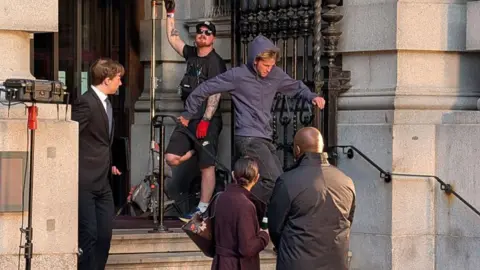 Conor McReynolds A man is running down the steps of a grey stone building. Past three people. A film crew member in the background is stood in the background.