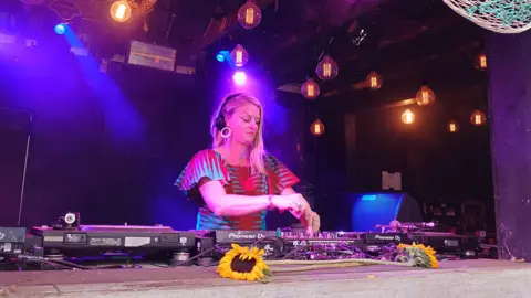 Faith Morton Faith Morton is standing in a DJ booth in a red dress with headphones on. She is looking down on a deck and pressing buttons. The lights are blue and purple.
