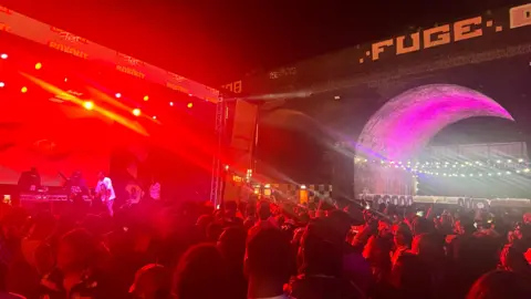 A crowd of people looking towards a stage that is lit up with red strobe lighting. They are at a festival which is taking place outdoors. The sky is dark and the crowd are standing under brick archways.