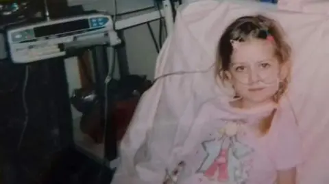 Sarah Gascoyne A young girl sits in a hospital bed, wearing a pink nightie with a cartoon fairy design on it. She has tubes attached to her face. She has brown hair and is smiling slightly. Hospital equipment can be seen in the background.