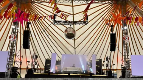 Shivani Chaudhari/BBC The DJ Stage inside a big tent. There are big speakers, a DJ booth in the middle with a screen behind it and a big disco ball above. There are various workers on the stage.