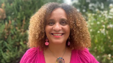 Susan Cole, who has tight curly brown hair, is wearing a pink top and pink dangly earrings in the shape of flowers and a colourful necklace. She is smiling at the camera.