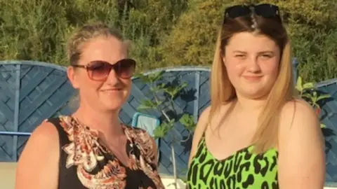 Tracy Marelli A family picture of Tracy and her daughter Sophie. They are both outside, posing for the photograph while smiling. Tracy is wearing a brown patterned dress, she has sunglasses on and has her curly hair tied up. Sophie is wearing a green and black patterned dress, her sunglasses are on her head, tying some of her long blonde hair back. Sophie is also smiling.