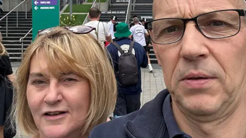 Handout A middle-aged man and woman take a selfie. In the background is a set of stairs and people milling around. 