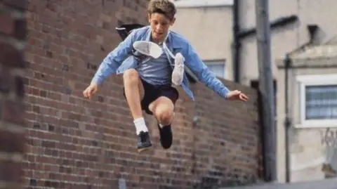 Working Title / BBC Films A scene from the film Billy Elliot shows him up in the air while he is mid jump on a street next to a brown brick wall. He is wearing black shorts and shoes, white socks and a blue shirt over a blue and white striped t-shirt. A pair of white trainers in hanging around his neck. 