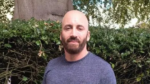 BBC A man in a grey T-Shirt is standing in front of a green hedge. He is bald and has a brown beard. He is smiling at the camera.