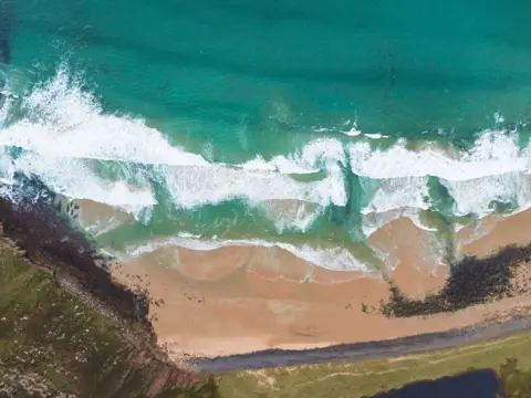 Getty Images High overhead view of Rackwick Bay on Hoy, showing pale sands, turquoise shallow waters, and gentle surf.