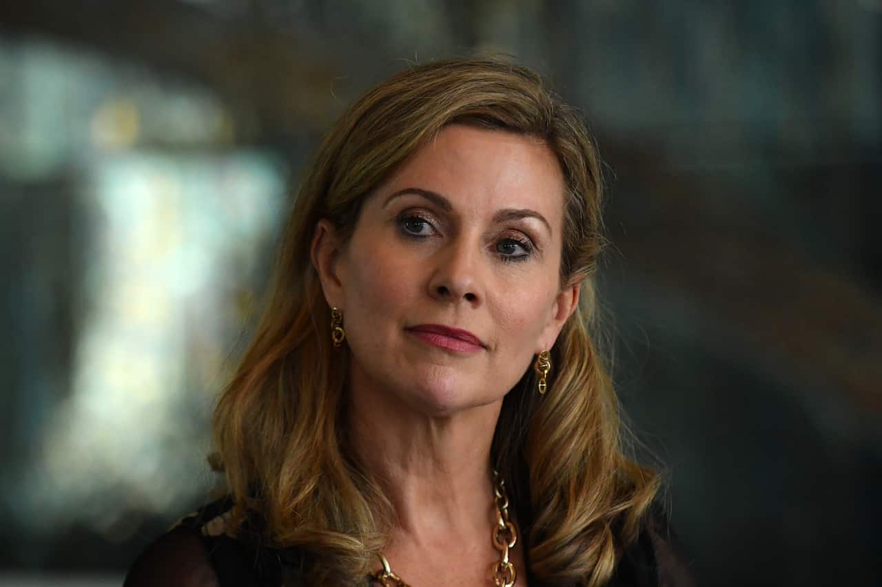 A head-and-shoulders shot shows a blonde woman with a serious expression and her head tilted slightly to the right. She is wearing a dark top, gold chain necklace, and small gold earrings.