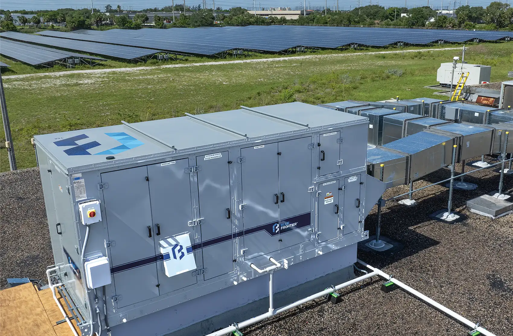 20250818-cooler-buildings-stronger-grid-new-approach-air-conditioning-built-in-energy-storage-riviera Photo of a large metal air conditioning unit with vents and access panels. The unit is adjacent to a field of photovoltaic panels.