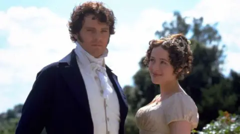 Getty Images Colin Firth and Jennifer Ehle pictured in costume together. Colin, on the left, wears a navy suit and a period white shirt. He has short curly brown hair. Jennifer, wears a beige, gold period dress and has brown curly hair which is tied up. She smiles whilst looking up at Colin Firth.