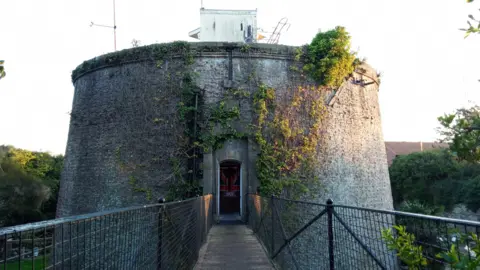 Owen Ward/BBC A cast-iron drawbridge leads towards the circular brick tower, which has ivy growing on part of its facade. A wooden door is at the end of the bridge.