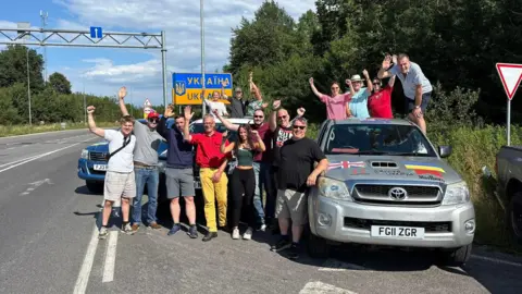 Driving Ukraine A group of about 20 people standing on the side of a road cheering, with a sign for Ukraine behind them. Alastair Caldwell is pictured with a group of three other people stood at the back of the Toyota Hilux pick-up.