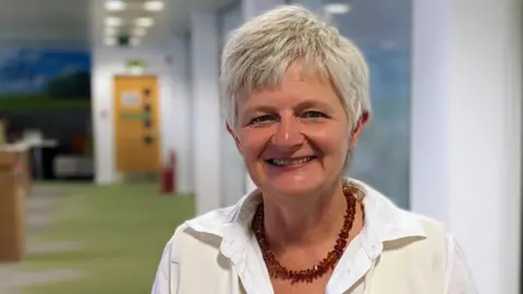 Age Scotland Katherine Crawford, a smart lady with short white hair smiles in her office which has scenic murals on the walls. She wears a white shirt and a red beaded necklace.