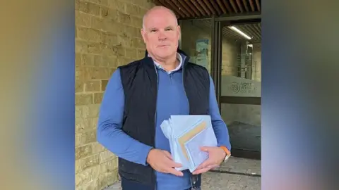 Stop Scotton Solar Farm A bald man wearing a light blue sweater and a dark blue gilet. He is holding a large white paper envelope and a bunch of forms and letters between his two hands.