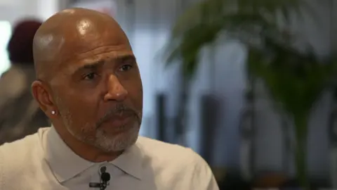 Marc Thompson, who has no hair and greying beard, is wearing a light coloured top with a collar and had a studded earring in his right ear.