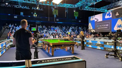 Tom Williams/BBC A snooker player is leaning on the table, taking aim for a shot. He is playing in a match at the Brentwood Centre. A referee wears all black as she stands behind him. In the foreground a camera operator films the action. In the background ar several rows of people in the crowd, all watching on.