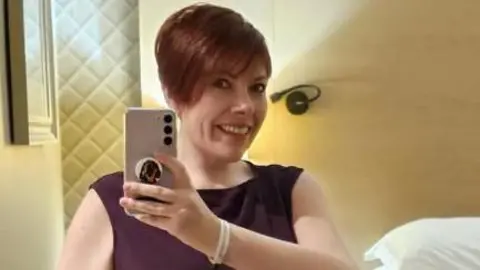 Kat Watson-Wood Kat Watson-Wood with short brown hair dressed in a burgundy evening dress in a hotel room holding her phone up to take a selfie. She is smiling.