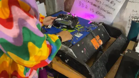 A woman standing next to a square blue robot.
She has a rainbow pattern on her sleeves and is holding a controller above the robot.