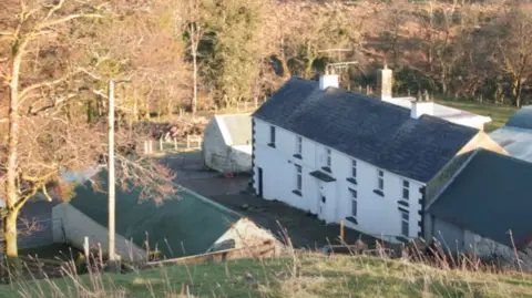 Frances Maguire The white farmhouse which has been in the McGoldrick family for generations is pictured from an aerial view. It is white and rectangular shape with black windowsills, cornerstones and a black roof. There is a white brick shed with a green roof, and there is another white shed. The house is surrounded by trees and green fields.