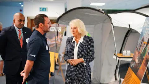 Ben Birchall/PA Wire Queen Camilla is given a tour by the Chief Executive Officer of ShelterBox, Sanj Srikanthan who is standing to the left dressed in a suit, during a visit to the headquarters of the charity.
She is wearing a dark dress with white polka dots and a white collar which extends vertically down the front of the dress. She is talking to a volunteer dressed in a blue polo shirt and stands in front of a tent.