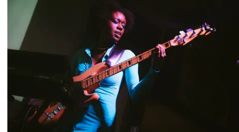 Saffron A woman in a blue jumpsuit is on a dark stage, playing the bass. Only her and her bass are lit up.