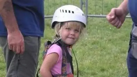 Angie Bunce-Mason Elana is in the middle looking towards the camera smiling. She has blonde hair and a fringe and is wearing a white helmet. She has a pink top on with a harness over the top.
