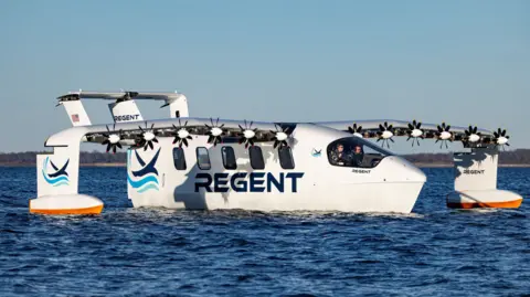 Amory Ross/Regent The prototype Seaglider is moving slowly across the surface of the sea on a sunny day. The machine looks like an aircraft with curved wings and six small propellers on each wing. It has hydrofoils on its undercarriage and wings. The machine is shown flying low over the surface of the sea. The fuselage is painted white and blue, and has five windows down its side and the word "Regent".