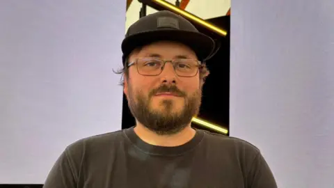 Shivani Chaudhari/BBC Joe Baker wearing a black sun cap and a black T-shirt and glasses. He has a black beard. He is smiling at the camera.