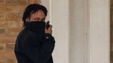 PA Media Larry Fraser, wearing a black coat he is pulling over his face to hide from the camera. He carries a phone and looks toward the photographer.